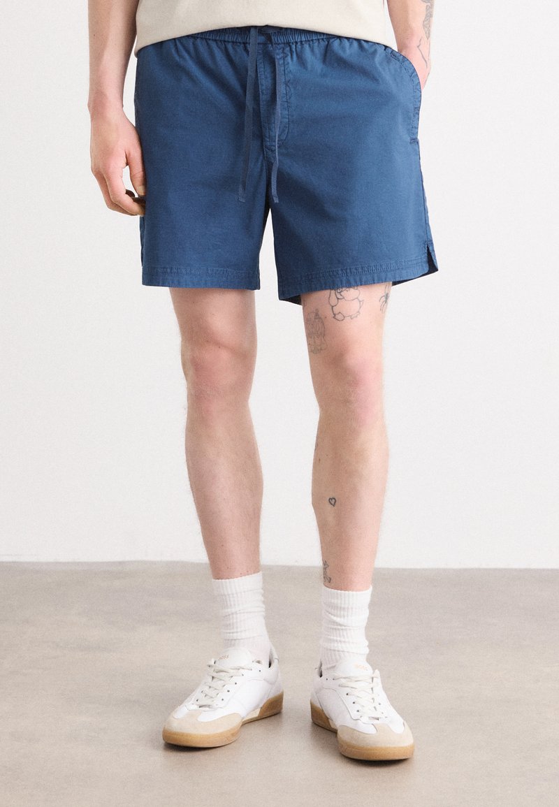 Shorts en coton bleu avec une taille élastique et un cordon de serrage. Comprend des poches latérales et une coupe droite, associés à des baskets blanches et des chaussettes.