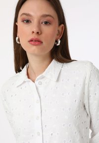 White blouse with floral embroidery, featuring cut-out patterns, a collared neckline, and button closure, paired with large silver earrings.