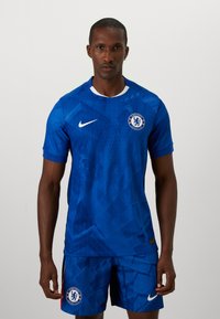Domowy strój Chelsea FC składający się z niebieskiej koszulki i spodenek, z geometrycznym wzorem, logo Nike oraz herb klubu na spodenkach, z białymi akcentami.