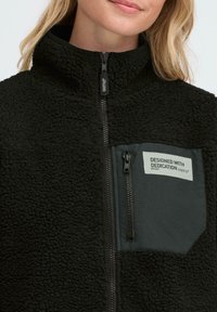 Black fleece jacket with high collar, zip closure, and a left chest pocket featuring a gray label with text. Soft, textured fabric.
