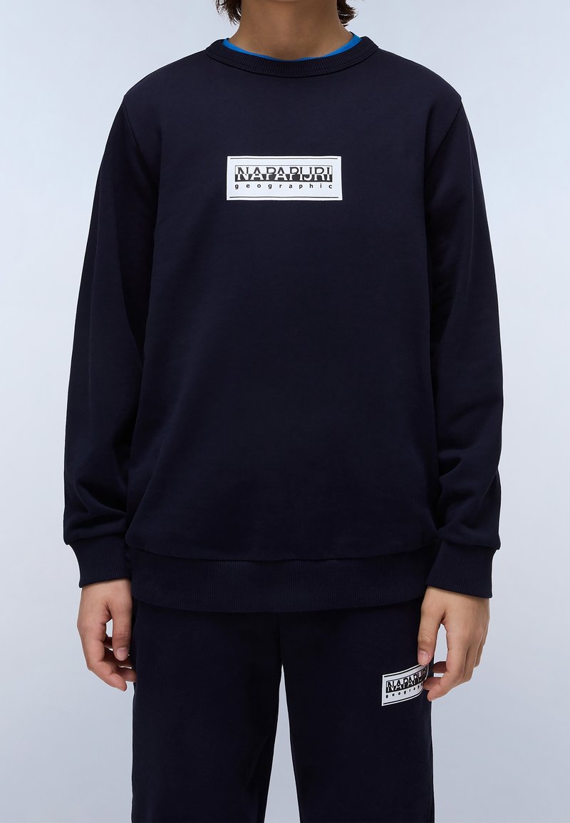 Personne portant un sweat-shirt noir et un pantalon, tous deux avec des logos Napapijri Geographic rectangulaires blancs au centre avant et à la cuisse gauche.