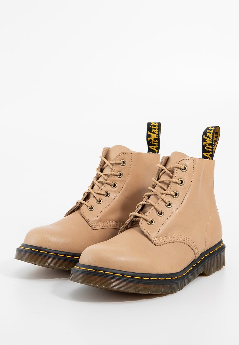 Botas de tobillo de cuero beige con cordones, costuras amarillas y suelas de goma negras. Presentan una lengüeta de tirón negra y amarilla en la parte trasera.