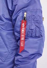 Chaqueta de nylon azul con un bolsillo con cremallera, que presenta una etiqueta de tela roja que dice "RETIRAR ANTES DEL VUELO." Textura suave y diseño compacto.