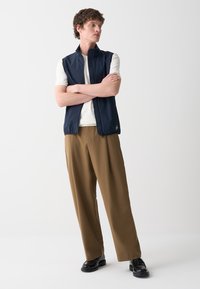 Gilet senza maniche blu navy con colletto alto e zip frontale, indossato sopra una maglietta bianca, abbinato a pantaloni marroni e scarpe nere.