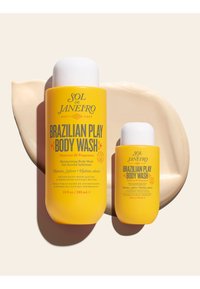 Dwie żółte butelki Sol de Janeiro Brazilian Play Body Wash z białymi zakrętkami na tle beżowej, kremowej plamy.