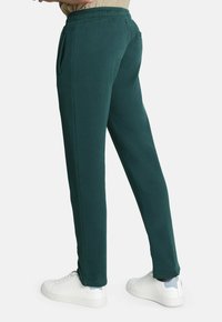 Pantalones de chándal verde oscuro con una cinturilla elástica, bolsillos laterales y piernas ajustadas, combinados con zapatillas blancas sobre un fondo neutro.