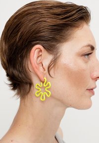 Žute polimerne naušnice u valovitom obliku cvijeta vise s uha, s glatkom teksturom i smjelim dizajnom, koje upotpunjuju kratku frizuru.