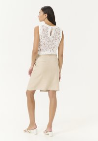 Top sin mangas de encaje con apertura en la espalda, combinado con una falda beige a la altura de la rodilla que cuenta con bolsillos traseros y una textura suave. Sandalias de tacón blancas.