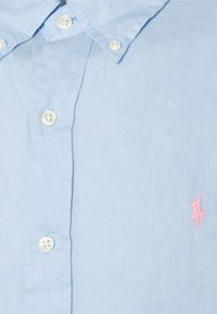 Světle modrá lněná košile s límečkem na knoflíky, s bílými knoflíky a růžovým vyšitým logem na levé straně hrudi.