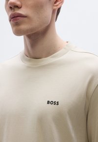 Bézs színű pamut póló kerek nyakkal, bal mellrészen fekete "BOSS" logóval. Sima textúra, minimalista dizájn.