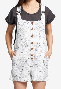 Salopette en coton blanc à motif floral avec des boutons en bois, matériel de sangle réglable en métal et poches avant. Portée par-dessus une chemise sombre.