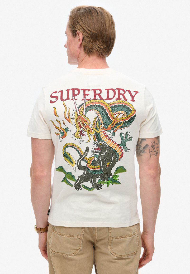 Camiseta de algodón blanca con un diseño gráfico colorido de un dragón y un gato negro, con "SUPERDRY" estampado arriba en rojo.