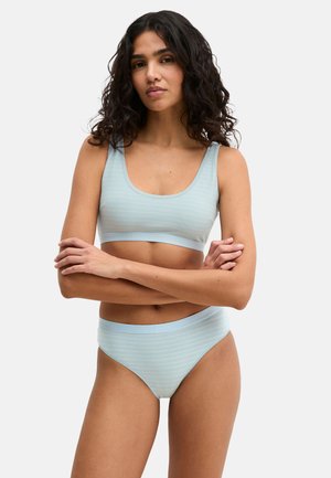 Vrouw met krullend haar, gekleed in een lichtblauwe gestreepte beha en bijpassend ondergoed, staand met gekruiste armen tegen een witte achtergrond.