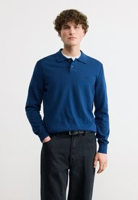 Νεαρός με κατσαρά μαλλιά που φοράει μπλε πουκάμισο πόλο με μακριά μανίκια και μαύρο παντελόνι, στέκεται με το ένα χέρι στην τσέπη, μπροστά σε λευκό φόντο.