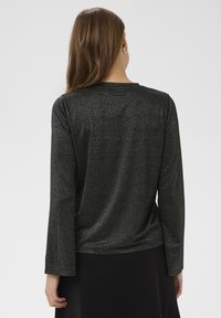 Žena s dlouhými hnědými vlasy, oblečená v třpytivém černém topu s dlouhým rukávem a černé sukni, zobrazená zezadu na jednobarevném pozadí.