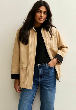 Veste beige avec un col, une poche avant et des poignets noirs ; associée à un pull noir et un jean bleu. Texture lisse, design décontracté.