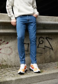 Jeans slim-fit azuis, material de algodão, combinados com sapatilhas esportivas brancas com detalhes em laranja e vermelho. Fundo cinza de concreto com grafite.