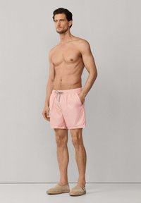 Homme torse nu portant un short de bain rose à motifs avec un cordon gris et des espadrilles beiges, debout devant un fond gris uni.