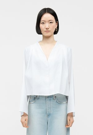 Donna con capelli neri corti, che indossa orecchini a cerchio dorati, una maglia bianca con scollo a V e jeans a vita alta color azzurro chiaro, in piedi davanti a uno sfondo semplice.