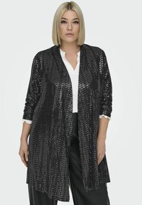 Mustris must kardigan, millel on pikk ja lahti istuv lõige, šaalikaelus ja kolmeveerandi pikkusega varrukad, kantud valge särk ja mustade pükstega.