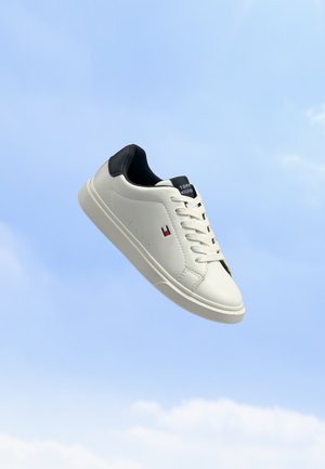 Λευκό sneaker χαμηλού ύφους με μαύρη φτέρνα, λευκά κορδόνια και μικρό κόκκινο και μπλε λογότυπο στο πλάι, με φόντο γαλάζιο ουρανό.