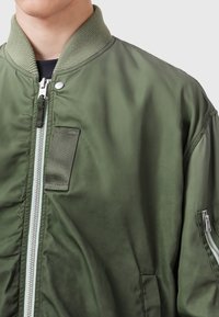 Green bomber jacket with a smooth texture, ribbed collar, zip closure, and a side pocket detail. Features a sturdy zip and snap button accents.
