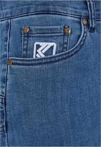 Sinine denimkangas sujuva tekstuuriga, mille esikotil on väike valge logo ja kuldsetest metallaktsentidest viimistletud õmblused.