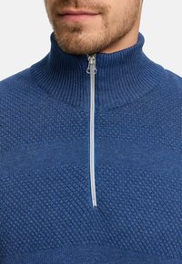 Blå zip-neck tröja med ett strukturerat stickat mönster och ribbad krage. Dragkedjan är silver och kontrasterar mot tygets färg och textur.
