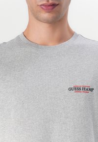 Tricou gri din bumbac cu guler rotund. Prezintă un logo brodat în culorile negru și roșu "GUESS JEANS" pe piept, cu "AMERICAN TRADITION" deasupra.