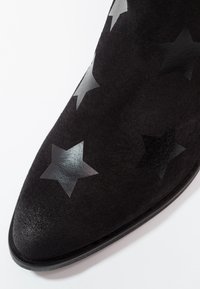 Stivale alla caviglia in suede nero con punta affilata e motivi a stella tono su tono in materiale lucido nella parte superiore. Suola liscia con dettagli minimi.