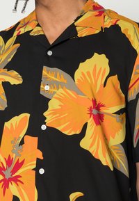 Korte mouwen overhemd met zwarte basis; heeft grote gele en oranje bloemmotieven en grijze bladeren; vijf witte knopen aan de voorkant.