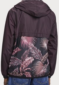 Person som bär en lila huva jacka med ett rosa och brunt tropiskt bladmönster på ryggen, i kombination med blå denimjeans.