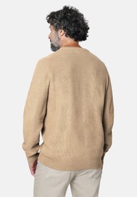 Hombre con cabello rizado oscuro y barba, vestido con un suéter de punto beige y pantalones claros, de espaldas ante un fondo liso.
