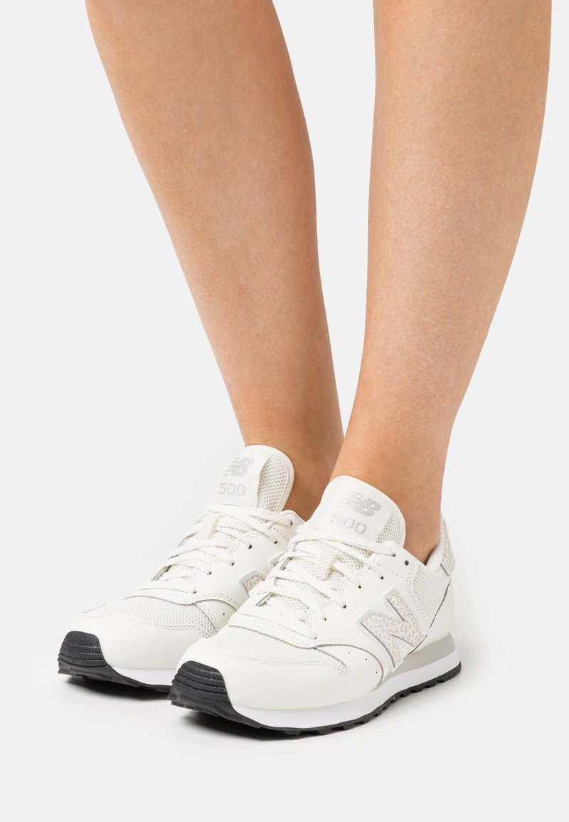 Paire de baskets blanches New Balance 500 avec empeignes en mesh et cuir portées sur des jambes nues, présentées sur un fond blanc uni.