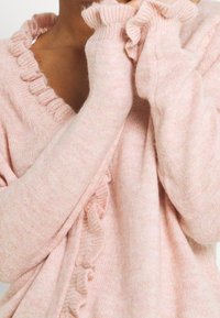 Měkký růžový pletený cardigan s volánkovými okraji límce a manžet, nošený osobou se zvednutýma rukama blízko obličeje.