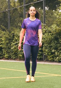 Violeta sporta krekls ar teksturētu rakstu, apvienots ar tumši ziliem legingiem ar rozā sānu akcentiem. Spilgti oranžas apavus. Zaļās zāles fons.
