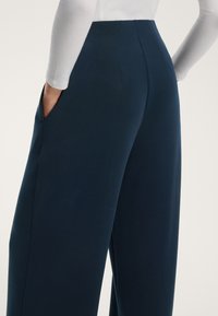 Široke modre hlače iz gladkega materiala z visokim pasom, ki imajo stranske žepke in subtilne šive.