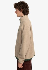 Beige fleece jacket with a high collar, green accents at the hem and zipper. Features a pocket on the left chest and textured fabric.