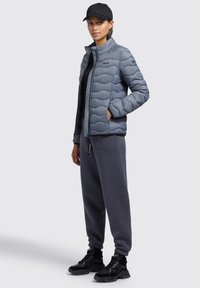 Lys grå polstret jakke med et struktureret design, sort lynlås, båret over en sort top og grå sweatpants. Sorte sneakers fuldender outfittet.