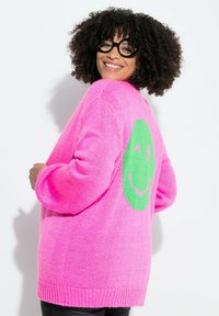 Maglione oversized rosa lavorato a maglia con un motivo a faccina sorridente verde sul retro. Presenta maniche lunghe e orlo a coste.