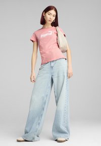 Rožinė medvilninė marškinė su balta logo, derinama su šviesiai mėlynomis plačiomis džinsinėmis kelnėmis. Modelis laiko šviesiai smėlio spalvos peties krepšį.