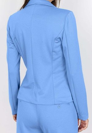 Femme portant un blazer ajusté bleu clair et un pantalon assorti, vue de dos sur un fond blanc uni.