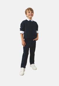 Granatowy sweter z białym kołnierzykiem, granatowe spodnie i białe sneakersy. Chłopiec stoi z jedną ręką w kieszeni, uśmiechając się, na jednolitym tle.