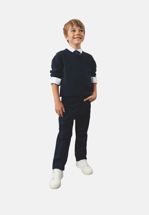 Granatowy sweter z białym kołnierzykiem, granatowe spodnie i białe sneakersy. Chłopiec stoi z jedną ręką w kieszeni, uśmiechając się, na jednolitym tle.