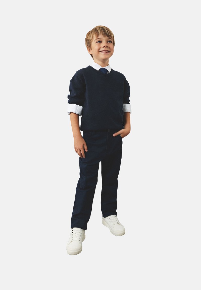 Granatowy sweter z białym kołnierzykiem, granatowe spodnie i białe sneakersy. Chłopiec stoi z jedną ręką w kieszeni, uśmiechając się, na jednolitym tle.