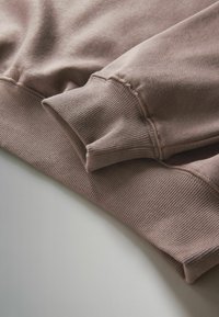 Hellbrauner, gerippter Stoff mit einer weichen Textur. Verfügt über ein umgeschlagenes Bündchen-Detail. Glatte Oberfläche mit Stichakzenten entlang der Kanten.