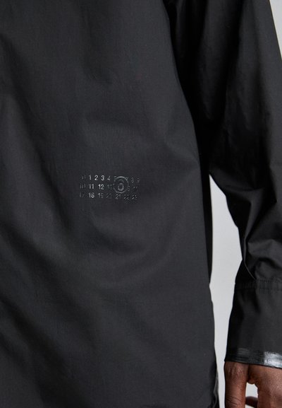 Sort vandtæt jakke med lange ærmer, der har et struktureret mønster af tal og en cirkulær detalje på venstre side nær kanten.
