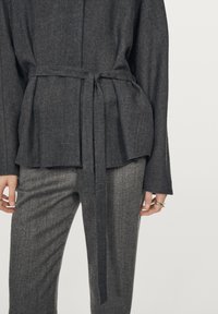 Top avvolgente grigio strutturato con cintura a fiocco, abbinato a pantaloni a righe grigie. Il tessuto sembra morbido con una vestibilità sartoriale e maniche lunghe.