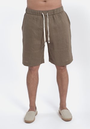 Uomo che indossa pantaloncini di lino marroni con coulisse e scarpe slip-on beige a righe, in piedi su sfondo bianco.
