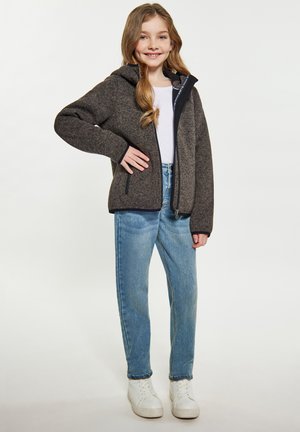 Giacca in pile marrone con cappuccio, cerniera nera e due tasche laterali, indossata sopra una maglietta bianca e abbinata a jeans blu e sneakers bianche.
